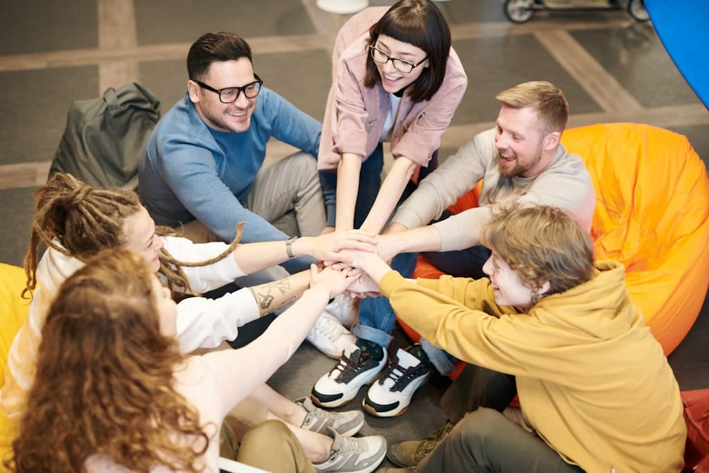 A diverse group of adults sit indoors, joyfully stacking hands together, symbolizing teamwork and camaraderie.